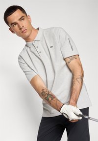 Lys grå polo skjorte med tre knapper, korte ærmer og brodert "CK"-logo på brystet. Har en sort "Calvin Klein" aksent på ærmet.