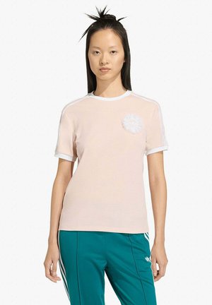 Maglietta rosa chiaro con bordi a coste bianchi sul collo e sulle maniche. Presenta una toppa circolare bianca con logo sul petto. Texture liscia.