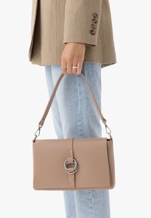 Ręka trzyma beżową skórzaną torebkę z detalem w postaci srebrnego pierścienia, w jasnoniebieskich dżinsach i brązowej marynarce z guzikami na rękawie.