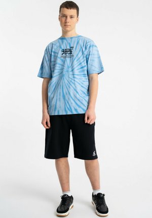 Młody mężczyzna stoi, ubrany w niebieski T-shirt typu tie-dye, czarne szorty i czarne sneakersy na białym tle.