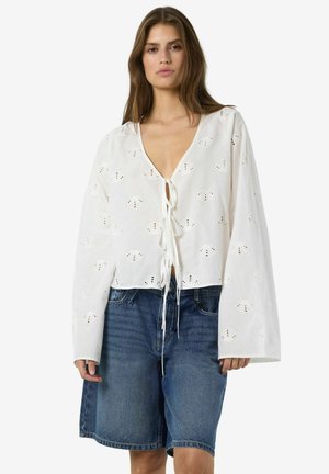 Ženska, oblečena v belo bluzo z izraženim očesnim vzorcem in vezalkami spredaj ter dolgimi rokavi, kombinirano z ohlapnimi modrimi boksaricami iz jeansa.