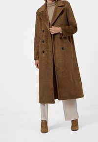 Brun, överstor, texturerad kappa med dubbelknäppning, svarta knappar och sidofickor, bärs över en beige turtleneck och kr ämfärgade byxor.