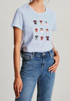 Personne portant un T-shirt rayé blanc et bleu avec des visages colorés de chiens portant des lunettes de soleil imprimés sur le devant, associé à un jean bleu.