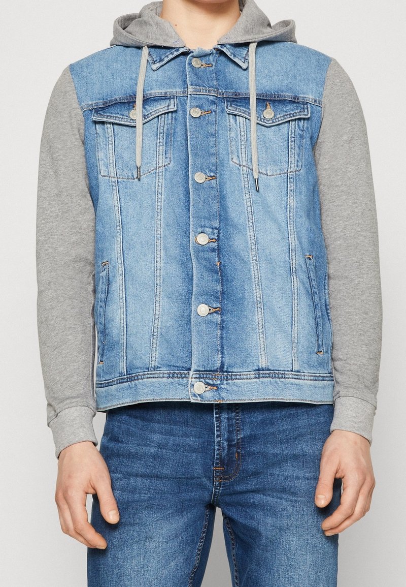 Jeansväst med grå huvtröjeärmar, ljusblå huvudtyg, två bröstfickor, silverfärgade knappstängningar och en lätt nött textur.