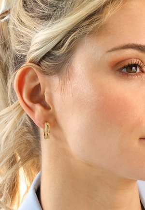 Goldener Creolen-Ohrring mit strukturiertem Design, verziert mit einem kleinen, klaren Stein. Wird am linken Ohr getragen und ergänzt den Hautton.