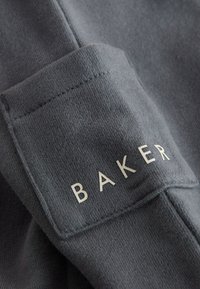 Gråt sweatshirtstof med en blød tekstur. Manchetten har ordet "BAKER" i hvide, fede store bogstaver. Simpelt design uden yderligere mønstre.