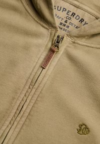 Gros plan sur un sweat à capuche beige zippé avec tirette de fermeture en cuir embossée "Superdry" et logo vert brodé sur le tissu.