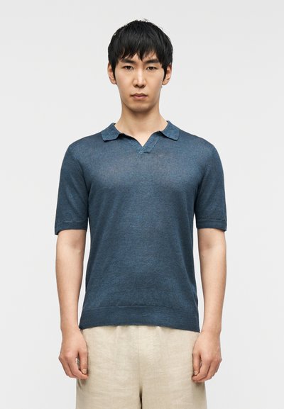 Polo bleu foncé, manches courtes, col en V, texture fine, coupe légèrement ample. Porté avec un short beige. Fond uni.