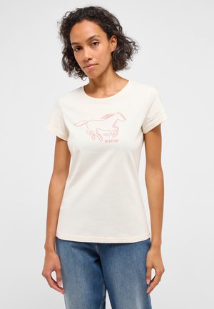 Woman with curly hair wearing a cream T-shirt with a red running horse graphic and "MUSTANG" text, paired with blue jeans.