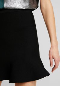 Svart ribbad stickad kjol med utsvängd nederkant; slät textur, figursydd design som sträcker sig just ovanför knäet, ofullständig vy av hand med neutral nagellack.