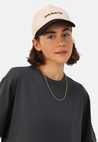 Casquette crème avec visière noire, texte "Club de lecture Zoom". Portée avec un t-shirt à manches courtes gris foncé et un collier en chaîne dorée.