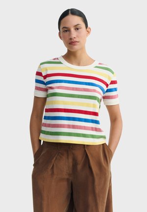 Young person with dark pulled-back hair wearing a short-sleeve t-shirt with multicolored horizontal stripes and brown trousers with hands in pockets.