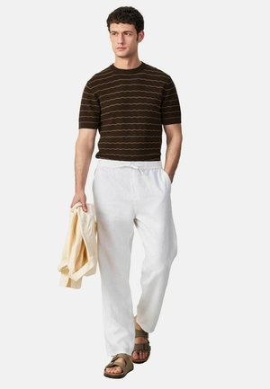 Man wearing a brown striped short-sleeve shirt, white drawstring pants, tan sandals, holding a beige jacket in one hand.