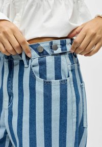 Mains attachant un bouton sur un pantalon en denim rayé verticalement bleu et bleu clair taille haute, portant un haut blanc et un bracelet argenté.