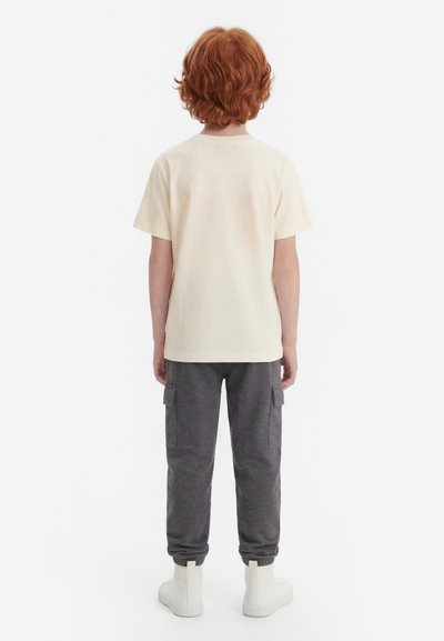 Vörös hajú gyermek háttal áll, krémszínű rövid ujjú pólót, sötétszürke cargo nadrágot és fehér tornacipőt visel, egyszerű fehér háttér előtt.