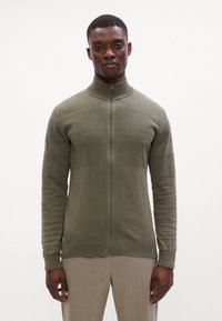 Giacca a zip verde oliva con colletto alto, design a pannelli testurizzati, polsini e orlo a coste. Realizzata in un tessuto morbido e elasticizzato.