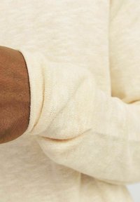 Suéter de color beige claro hecho de una suave tela texturizada. Detalle de puños acanalados visibles y ajuste ligeramente holgado alrededor de la muñeca.