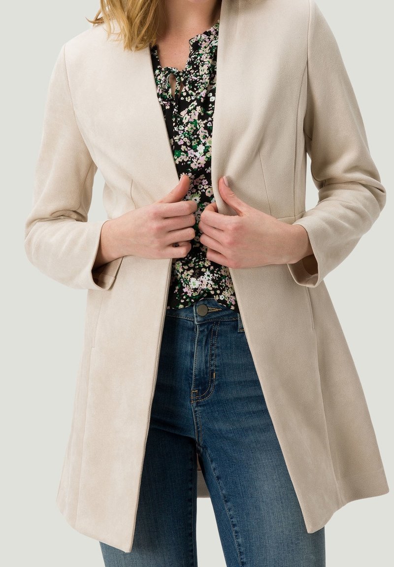 Femme portant un long blazer beige, un chemisier fleuri et un jean bleu, tenant les revers du blazer avec les deux mains.