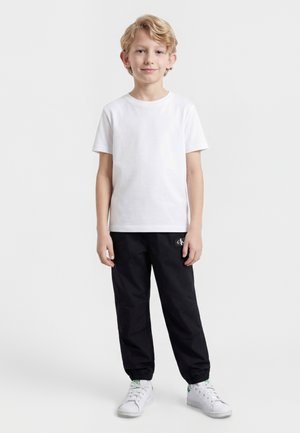 Blonder Junge steht, trägt ein schlichtes weißes T-Shirt, schwarze Jogginghosen und weiße Turnschuhe vor einem schlichten hellen Hintergrund.