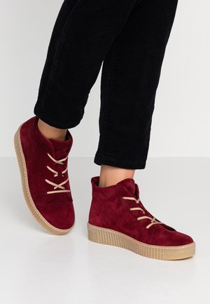 Person iført røde ruskindshøje sneakers med beige snørebånd og tykke beige såler, kombineret med sorte fløjlsbukser.