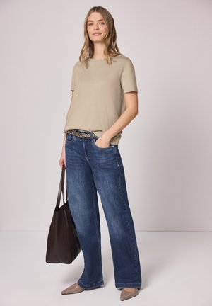 Kobieta stoi z jedną ręką w kieszeni, ubrana w beżowy t-shirt, niebieskie szerokie dżinsy, pasek w panterkę, beżowe szpilki, trzyma ciemnobrązową torbę na ramię.