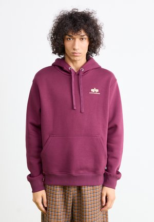 En ung person med krøllet hår har en burgunder Alpha Industries hettegenser og brune rutete bukser, stående mot en enkel hvit bakgrunn.