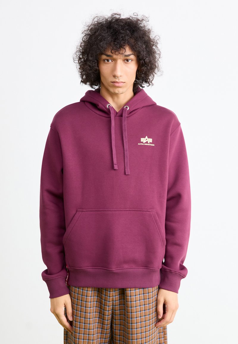 Persona joven con cabello rizado que lleva una sudadera color burdeos de Alpha Industries y pantalones de cuadros marrones, de pie contra un fondo blanco liso.