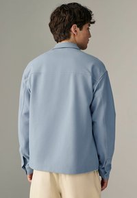 Camisa de manga larga azul claro con cuello, que presenta costuras visibles y un ajuste holgado, mostrada desde la parte posterior sobre un fondo neutro.