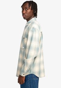 Cămașă cu model tartan în nuanțe de albastru deschis și crem, cu mâneci lungi, un buzunar pe partea stângă și o croială lejeră. Material moale, texturat.
