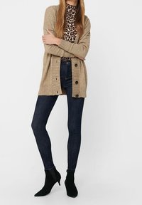 Nainen, jolla on päällään beige nappikauluspaita, leopardikuosiset paita, tummat kapeat farkut ja mustat nilkkurit, seisoo kädet ristissä.