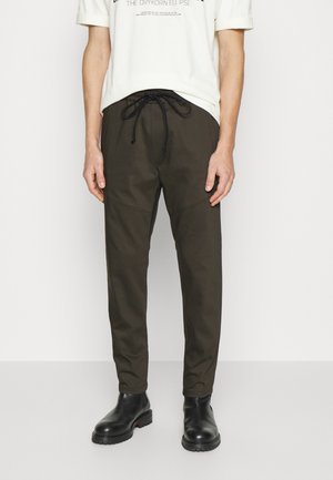 Mann trägt dunkelolivgrüne Kordelhose, schwarze Stiefel und ein weißes T-Shirt mit hochgekrempelten Ärmeln vor schlichtem weißem Hintergrund.