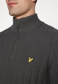 Schiefergrauer Strickpullover mit geripptem Design, Halbreißverschluss-Kragen und gelb gesticktem Logo auf der Brust. Strukturierte Stoffoberfläche.