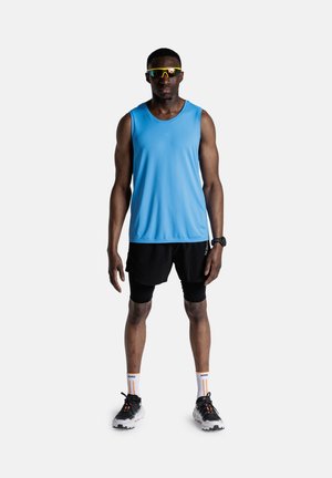 Man draagt een blauw mouwloos shirt, zwarte shorts, zwarte compressietights, hardloopschoenen, reflecterende zonnebril en sporthorloge, staat.