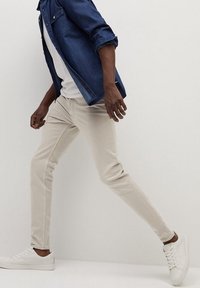 Pantalones ajustados de color beige claro con una textura suave, combinados con una camiseta blanca y una camisa de mezclilla, acompañados de zapatillas blancas.