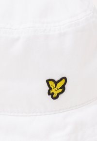 Bijela kapica od pamuka s šivanim šavovima. Ima mali izvezeni logo insekta u crnoj i žutoj boji. Glatka tekstura.