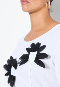 Maglietta bianca con applicazioni di fiori in tessuto nero e accenti quadrati bianchi. Presenta una texture liscia e un colletto rotondo.