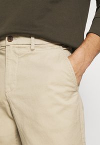 Chinos bej din bumbac cu buzunare laterale și un nasture maro la talie, asortați cu o cămașă verde cu mânecă lungă, textura materialului fiind vizibilă.