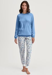 Lichtblauw overhemd met lange mouwen gecombineerd met witte pyjamabroek met een bloemenpatroon in blauw, groen en zwart. Elastische manchetten aan de broek.