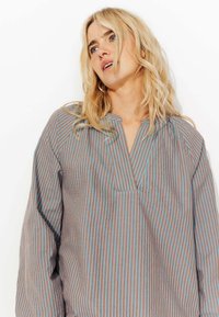 Blouse rayée avec des bandes verticales bleues et marron, décolleté en V et tissu doux avec une coupe décontractée. Manches longues avec détails froncés.