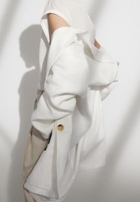 Osoba w białej bezrękawnikowej bluzce i beżowych spodniach, trzymająca białą kurtkę zawieszoną na ramieniu na tle jasnym.