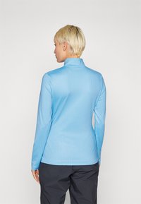 Maglietta azzurro chiaro a maniche lunghe con colletto alto, caratterizzata da un motivo testurizzato, forma aderente e vita nera a contrasto, indossata con pantaloni scuri.