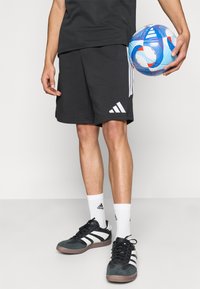 Μαύρο αθλητικό σορτς με λευκές ρίγες Adidas, σε συνδυασμό με μαύρα και γκρι παπούτσια και λευκές κάλτσες. Μια πολύχρωμη ποδοσφαιρική μπάλα κρατιέται στο ένα χέρι.