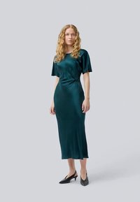 Vestido midi de cetim verde escuro com mangas curtas, detalhe de cintura torcida e uma textura lisa e suave. Combinado com sapatos de salto preto de bico afiado.