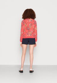 Camisa larga de manga larga con estampado floral rojo, acentos en azul y amarillo, combinada con shorts marinos y sandalias blancas. Vista trasera, tejido suave y corte relajado.
