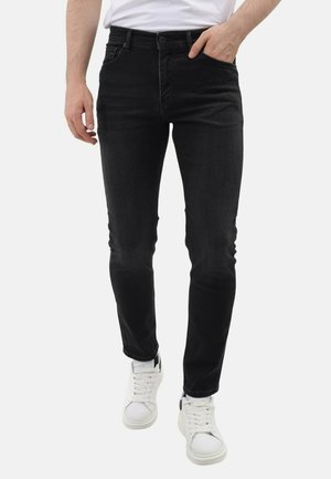 Persoon draagt slimfit zwarte spijkerbroek en witte sneakers, met één hand in de zak, staand tegen een effen lichte achtergrond.