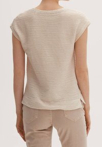 Helles beige kurzärmliges Oberteil mit einem strukturierten, wellenförmigen Muster. Weicher, lässiger Schnitt mit einem runden Halsausschnitt und leichten seitlichen Schlitzen am Saum.