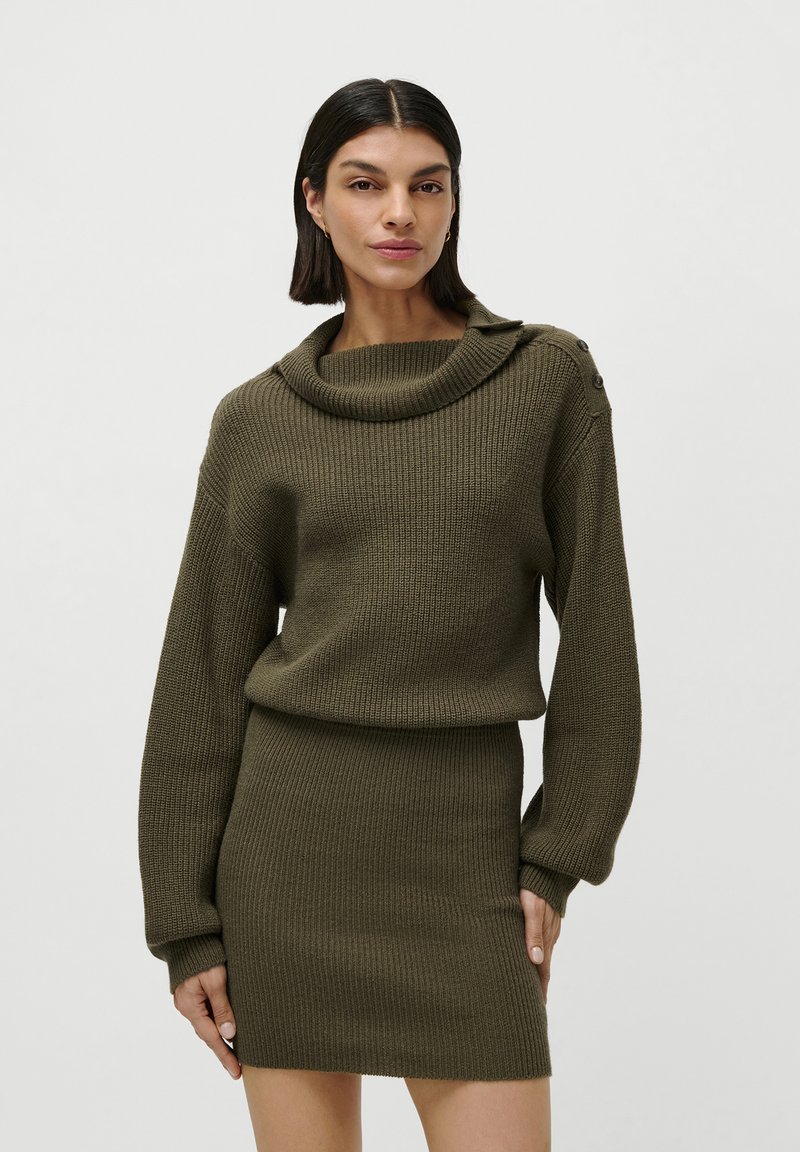 Donna che indossa un maglione a coste di un verde oliva e una gonna coordinata, in piedi con un'espressione neutra contro uno sfondo semplice.