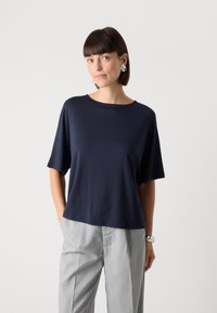 Top navy blue a maniche corte realizzato in tessuto morbido con una vestibilità rilassata. Collo rotondo, texture liscia, abbinato a pantaloni a strisce grigio chiaro.