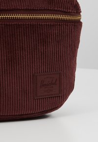 Bordowa torba z materiału kordurowego z poziomym złotym zamkiem błyskawicznym i naszytą metką marki Herschel na przodzie.