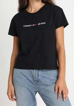 Kobieta w czarnej koszulce Tommy Jeans z logo w kolorze białym i czerwonym, w połączeniu z jasnoniebieskimi dżinsami z wysokim stanem, stojąca na jednolitym tle.